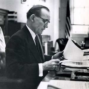 White man in suit and tie and glasses sitting at desk looking at papers