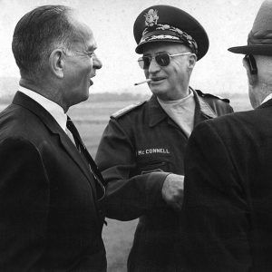 White man in military garb and a cigarette sticking out of his mouth standing between two white men in suits