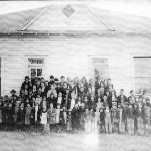 Large group of people standing before single story white wooden building