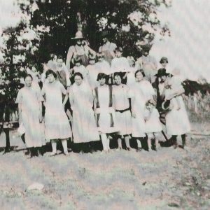 Group of white children and adults standing next to single story building and mules and wagon