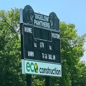 Metal scoreboard saying "Bigelow Panthers" on top with trees in background