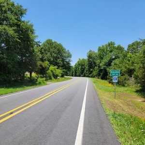 Rural highway with trees on either side and green sign saying "Houston population one hundred forty-three"