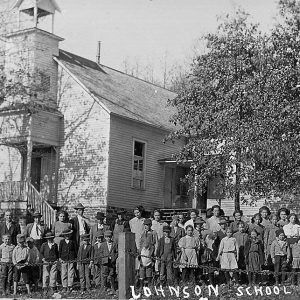 Large group of white children and white adults standing in front of multistory wooden building with bell tower