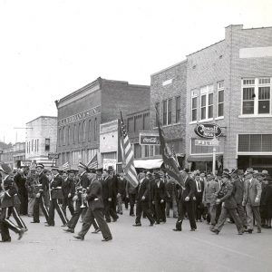 Uniformed white boys and men some with instruments and flags parading down street lined with multistory storefront buildings