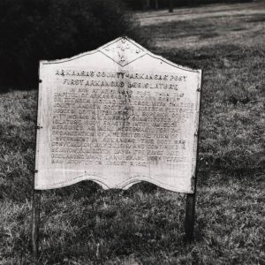 Metal sign leaning to the right with words at the top saying "Arkansas County Arkansas Post First Arkansas Legislature"