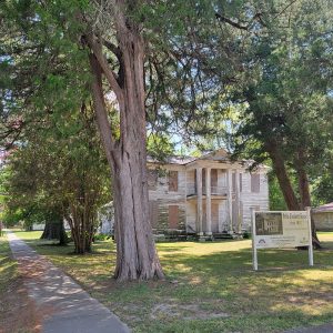 Multistory white wooden house with chipped paint and boards on windows and tall trees around it and informational sign