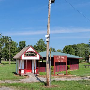 Single story red and white wooden building next to pavilion with park area in background