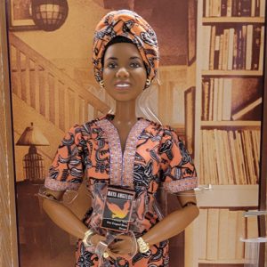 Doll of African American woman in black and orange flowery dress and head covering holding a book in front of her