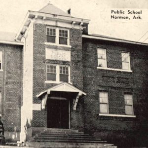 Multistory brick building with covered entrance and large bell in front on a pedestal