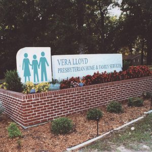 White signs saying "Vera Lloyd Presbyterian Family Services" and picturing three people holding hands atop red brick planter