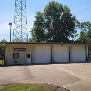 Single story beige metal building with three garage doors and a tower behind it