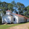 Single story white wooden church building with trees behind