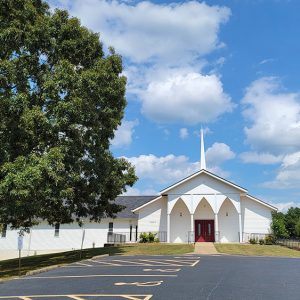 Multistory white wooden church building with steeple and parking lot