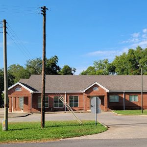 Single story red brick post office building with parking lot and trees behind