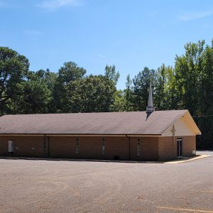 Single story red brick church building with steeple and large parking lot and trees in background