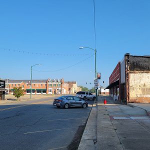 Single story storefront buildings along road with multistory orange brick building in distance