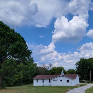 Single story white wooden church building with steeple and gravel road and trees in background