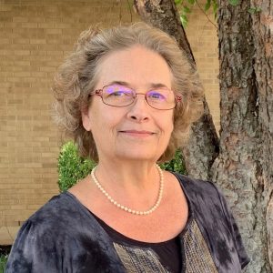 White woman wearing glasses and pearls standing in front of a tree and a brick wall