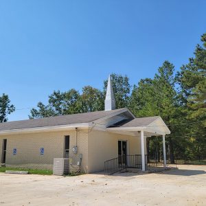 Single story blond brick church building with covered entrance and steeple