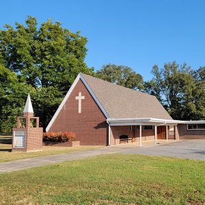 red brick church building with brick sign holder with a pointed tower on top and parking lot