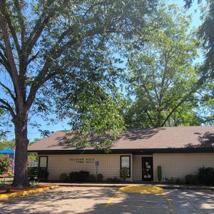 Single story tan concrete block building with "Sulphur Rock Town Hall" on the front with parking lot and overhanging trees