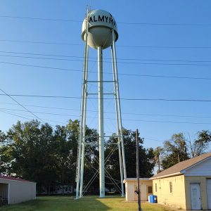 Metal cylindrical water tank on tall legs with "Almyra" in large black letters