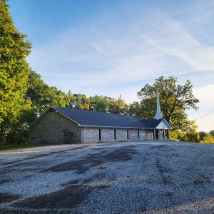 Single story tan brick church building with white spire with parking lot and trees in background