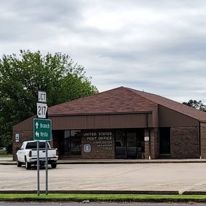 Single story brown brick post office building with parking lot