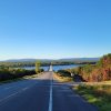 Country road heading toward bridge over river with hills in the distance and trees and both sides of road