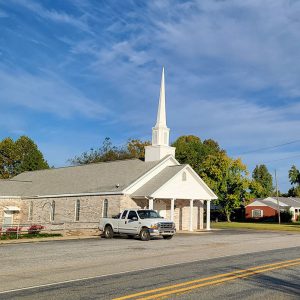 Single story blond brick church building with steeple and covered entrance supported by columns