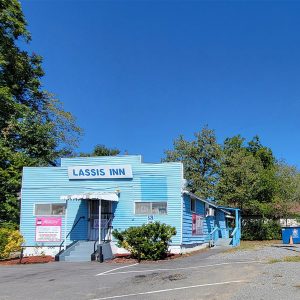 Single story blue wooden building with parking lot and trees