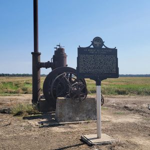 Rusted machinery on a concrete block and a historical sign in the middle of a field