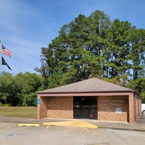 Single story orange brick post office building with parking lot and trees in background