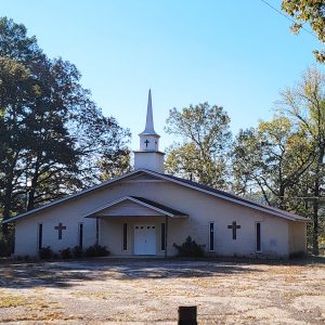 Single story white brick church building with white steeple and parking lot with trees behind building