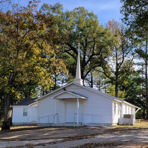 Single story white wooden church building with steeple and trees in background