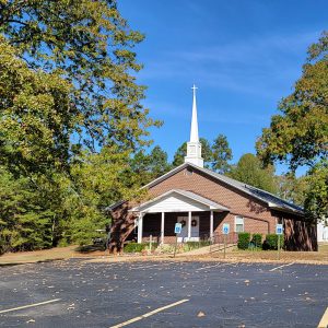 Single story red brick church building with white steeple and parking lot