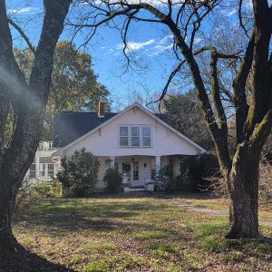 Two-story white wooden house with porch and huge trees in the yard