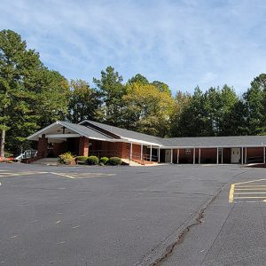 Single story red brick church building with parking lot and trees in background