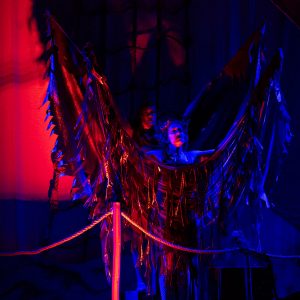 Two actors holding up fabric that has been ripped to shreds on a stage lit in red and blue