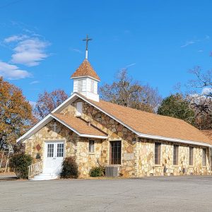 Single story rock church building with small steeple with cross on top and parking lot