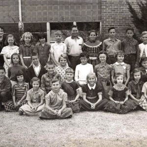 White children and an older woman standing and sitting in front of a building