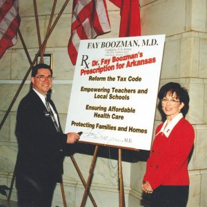 White man and white woman standing at a campaign sign saying "Dr. Fay Boozman's Prescription for Arkansas"