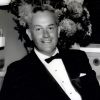 White man in a tuxedo and bow tie with flowers behind him