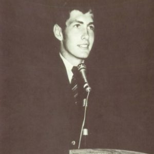 White young man in with short hair dressed in suit and standing at microphone