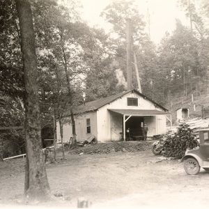 Man standing on porch of house in wooded area with cars