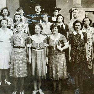 Group of white women in dresses standing around a white man in priest's garb