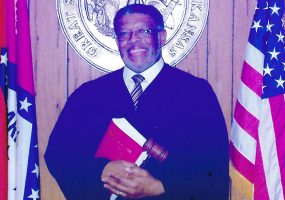 African American man in judicial garb standing between state and national flags