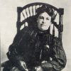 White woman in dark striped dress with a high collar sitting in rocking chair