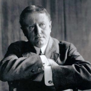 Seated white man in suit with arms crossed across back of chair