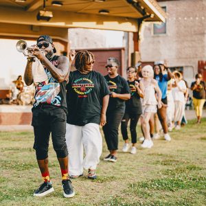 Group of people of various races dancing in a line behind an African American man playing trumpet